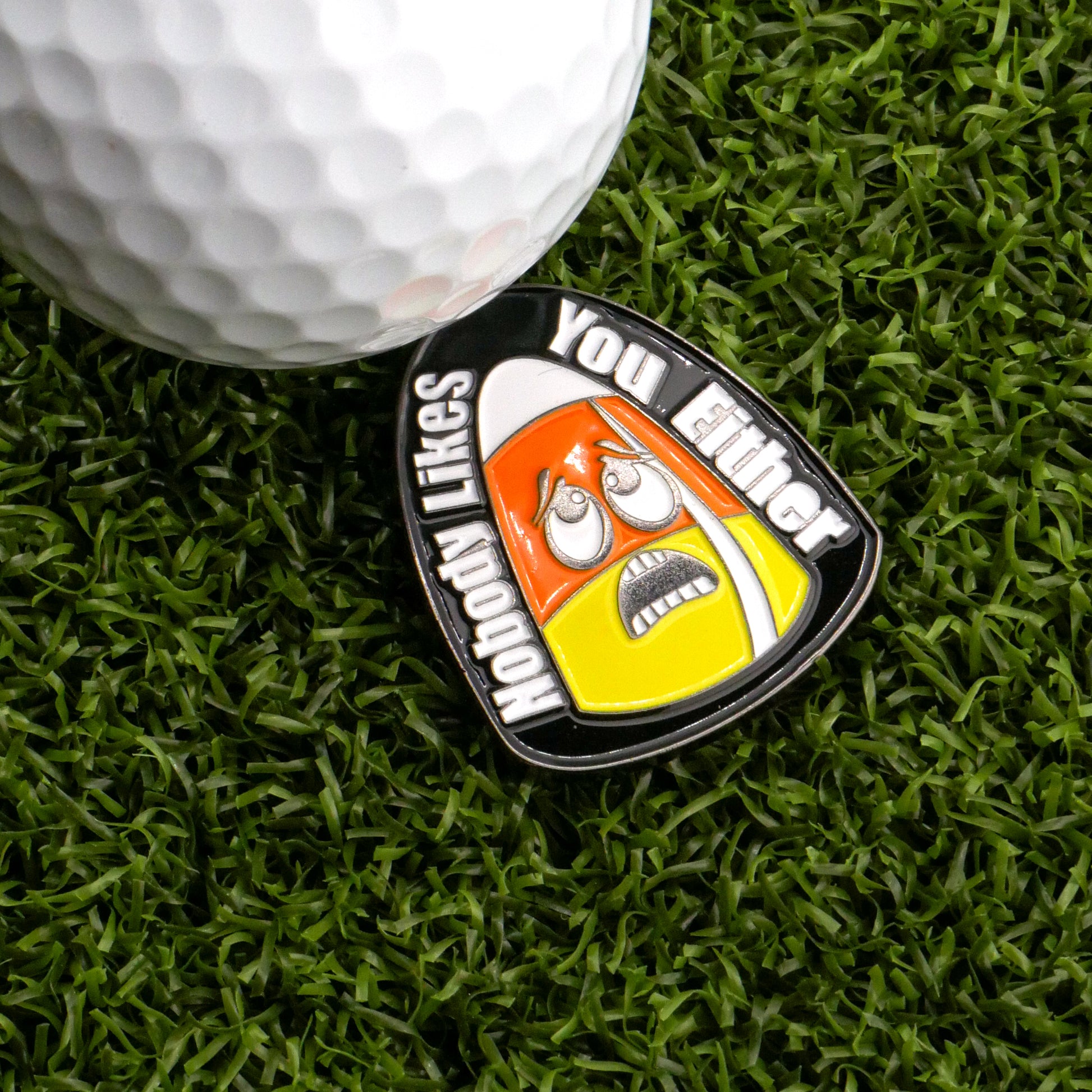 A golf ball on artificial grass sits next to the Sad Candy Corn Ball Marker from Ballmarkers.com, shaped like candy corn with an anxious face and the words "Nobody Likes You Either.