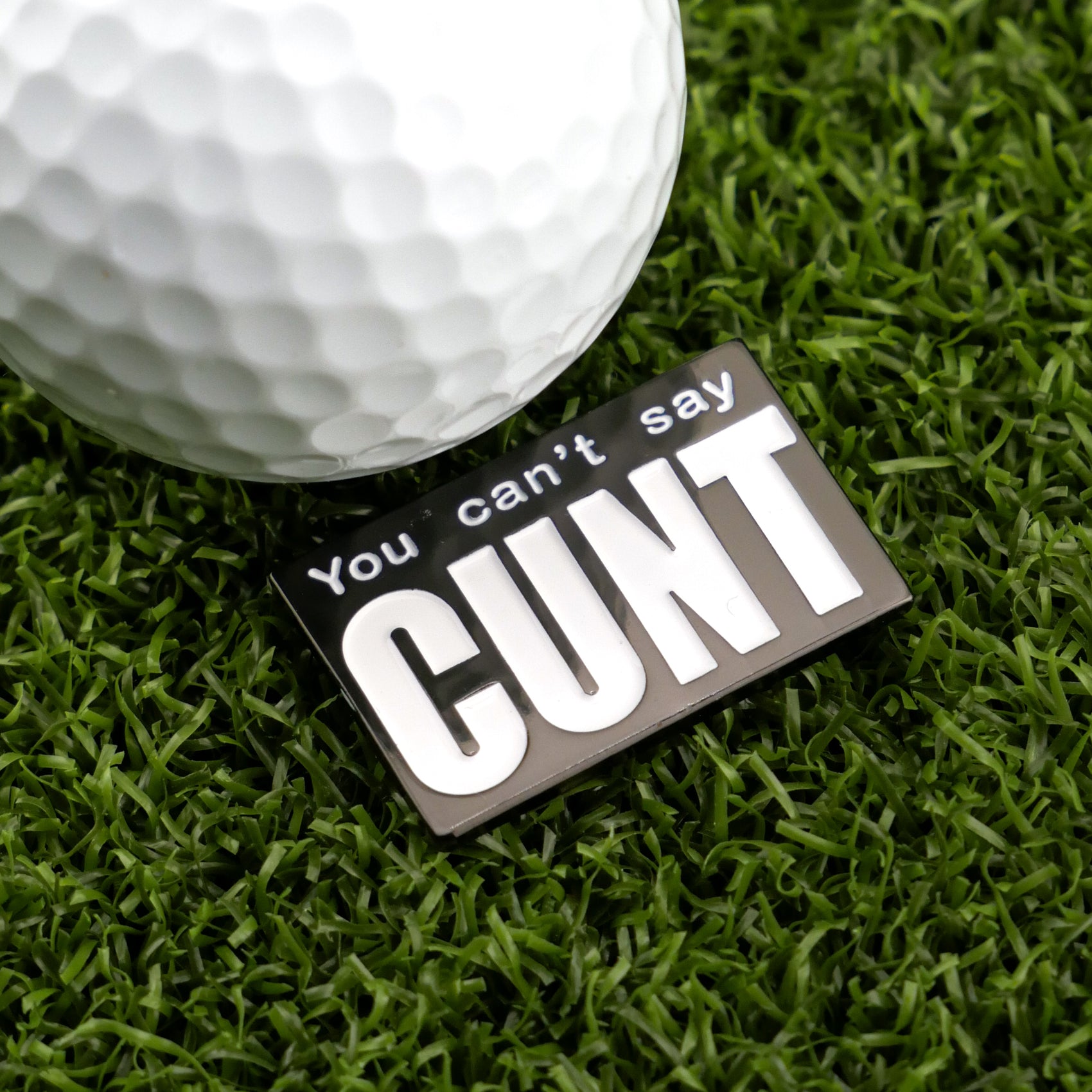 A Logie Joe's Golf ball and marker rest on artificial grass next to a black and white "You Can’t Say Cunt" sign—an ideal humorous gift from Logie Joe's Golf for those who love quirky golf accessories.