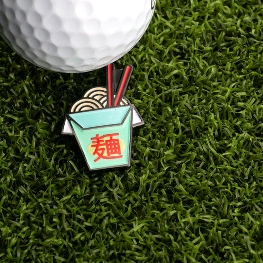 A close-up of a golf ball on artificial grass beside Logie Joe's Golf Chinese Take-Out Ball Marker, shaped like an enamel takeout box with chopsticks and a red Chinese character.
