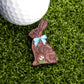 The Chocolate Bunny Ball Marker by Ballmarkers.com, shaped like a bunny with a blue bow and flower, sits on green turf beside a white golf ball—an adorable Easter golf accessory for the course.