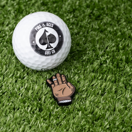 A Pins and Aces golf ball rests on artificial grass beside the Chubs Hand Ball Marker, a magnetic marker shaped like a cartoon peace sign hand, crafted with vibrant enamel paint for extra style.
