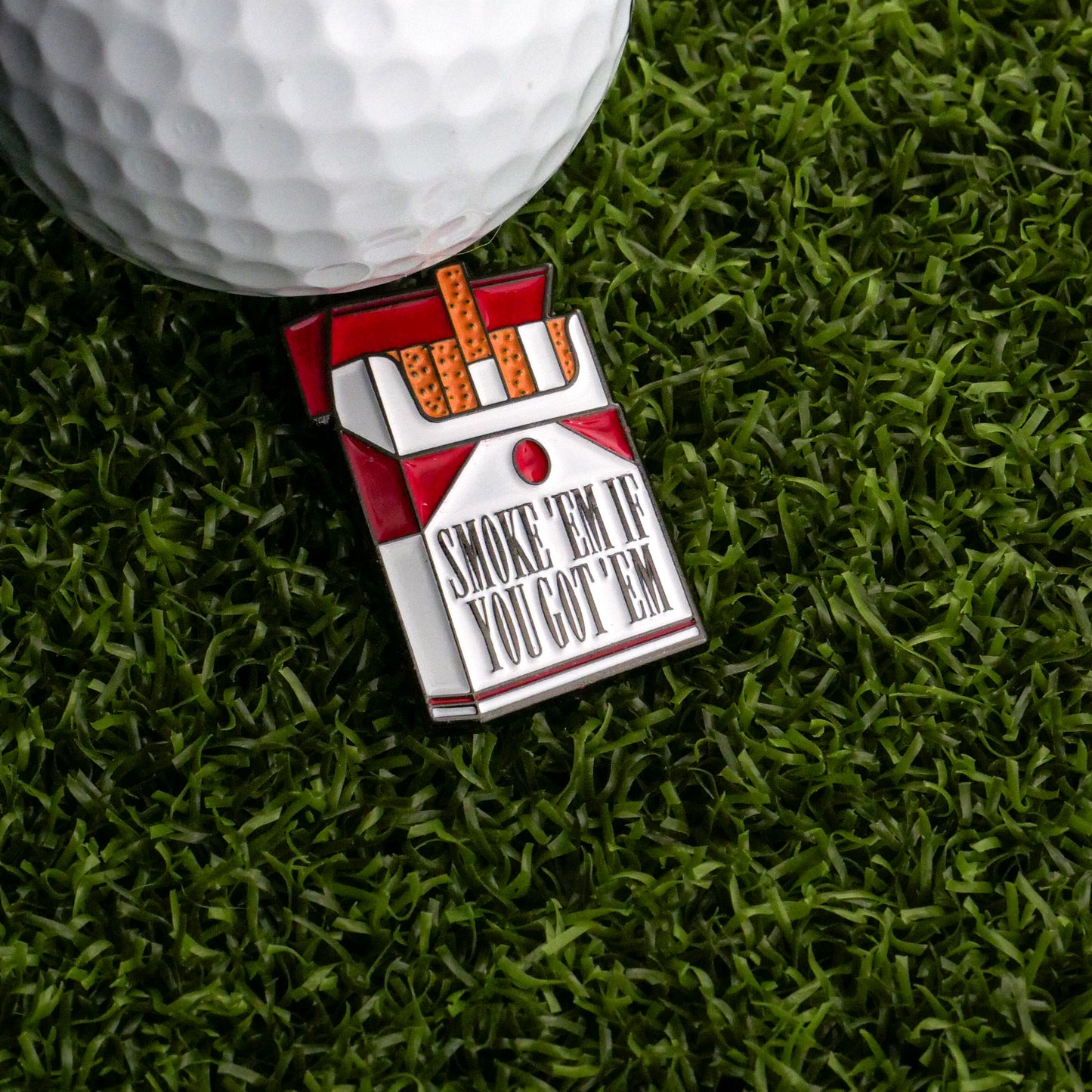 A white golf ball rests on artificial grass next to the Ballmarkers.com Cigarette Pack Ball Marker, featuring "SMOKE ‘EM IF YOU GOT ‘EM"—a quirky accessory for your next round.