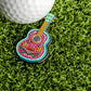 The Cinco De Mayo Guitar Ball Marker by Ballmarkers.com features colorful Mexican folk art floral designs and sits on green artificial grass near a white golf ball—perfect as a festive, unique golf accessory.