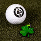 A white Pins and Aces golf ball sits on green turf beside a Shamrock Ball Marker by Pins and Aces, shaped like a four-leaf clover with a yellow flag.