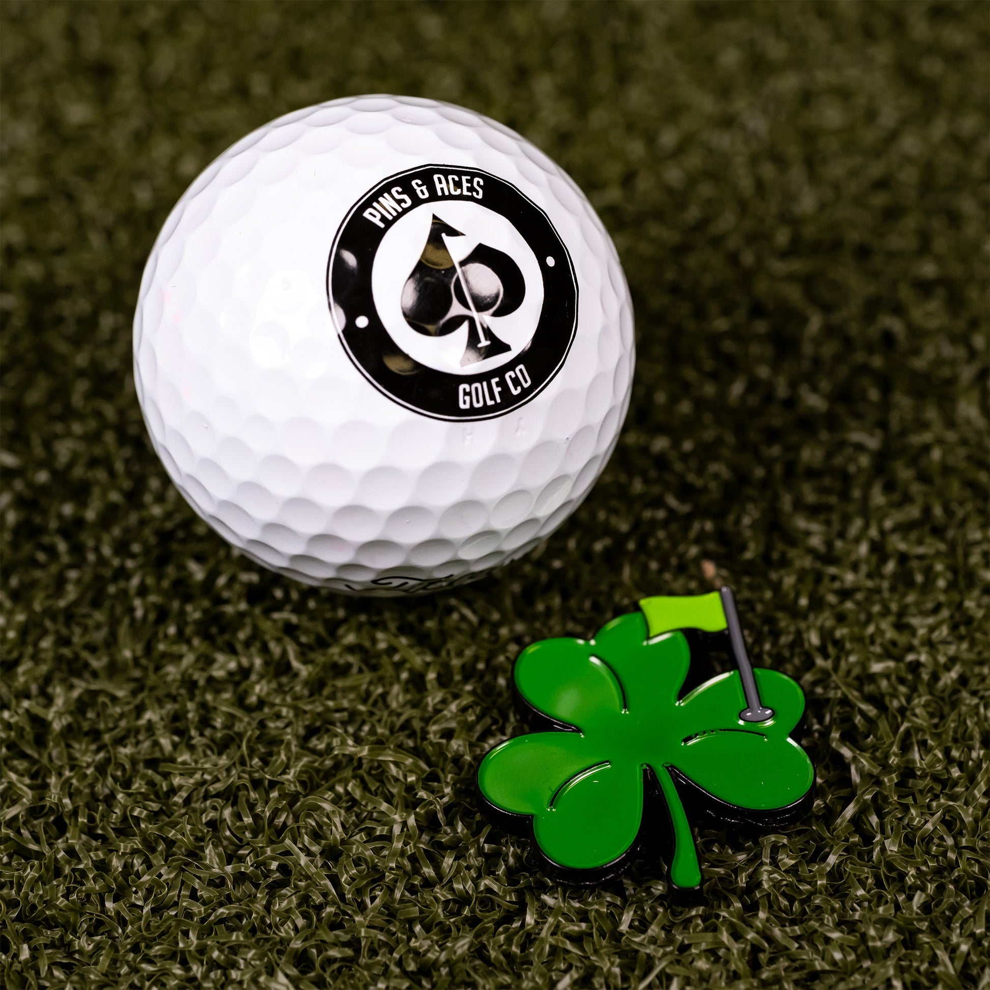 A white Pins and Aces golf ball sits on green turf beside a Shamrock Ball Marker by Pins and Aces, shaped like a four-leaf clover with a yellow flag.