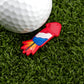 A golf ball sits on artificial green grass beside Logie Joe's Golf Cock Rocket, a colorful, rocket-shaped golf ball marker with red, white, blue, yellow, and orange details—one of the most humorous golf accessories.