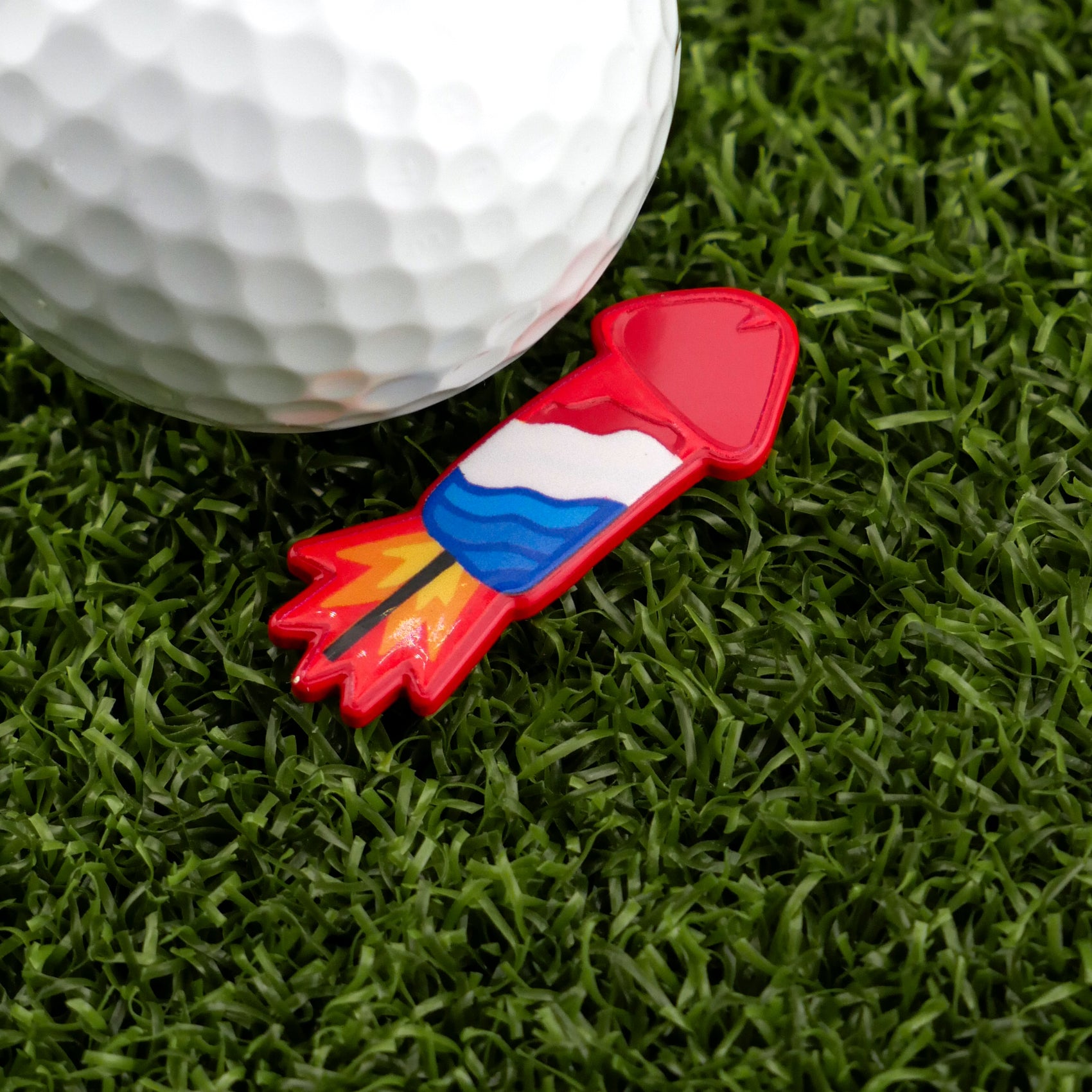 A golf ball sits on artificial green grass beside Logie Joe's Golf Cock Rocket, a colorful, rocket-shaped golf ball marker with red, white, blue, yellow, and orange details—one of the most humorous golf accessories.