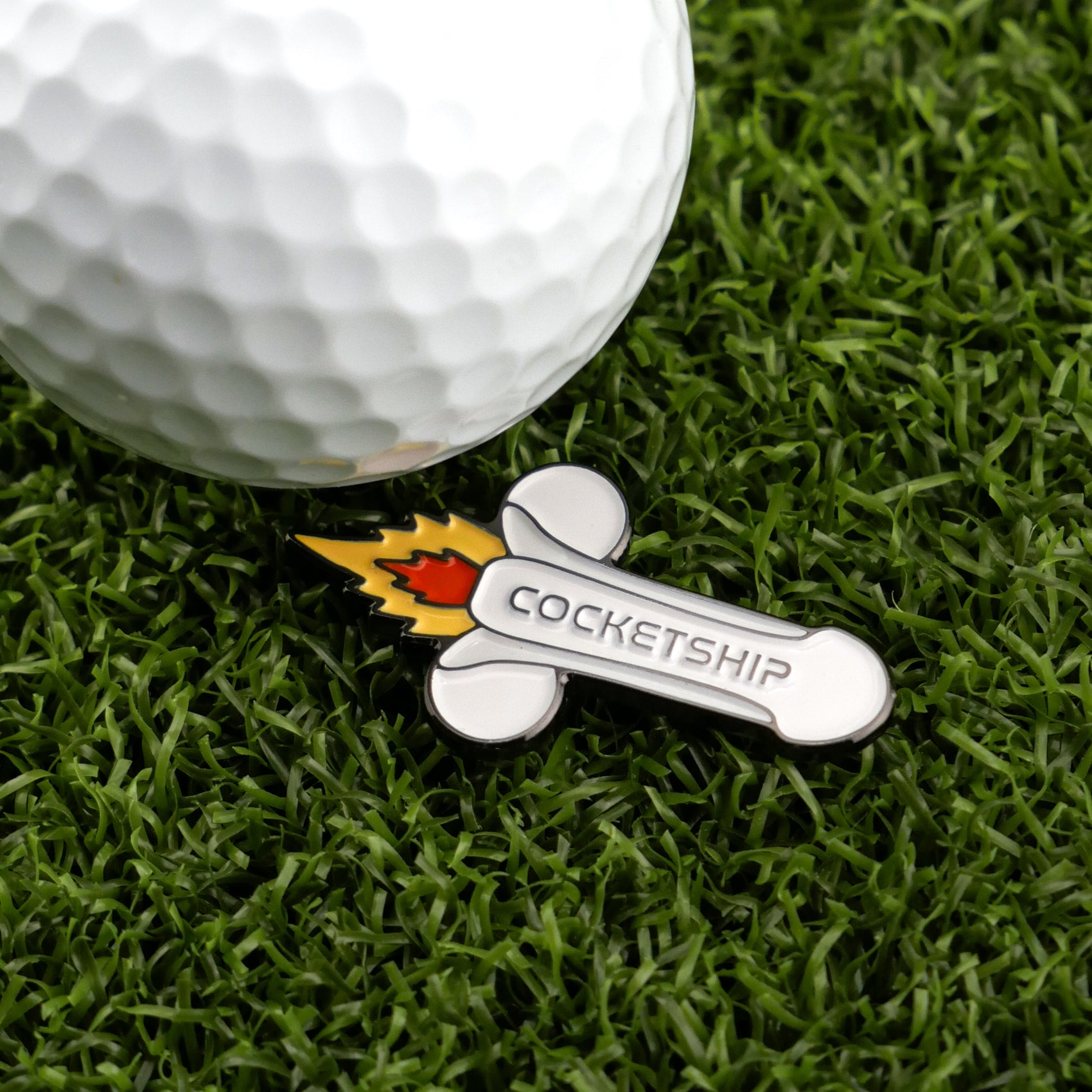 A golf ball on artificial grass sits beside Logie Joe's Golf Cocketship—a cartoon rocket-shaped, cheeky ball marker that adds playful humor to your game with its unique phallic design.
