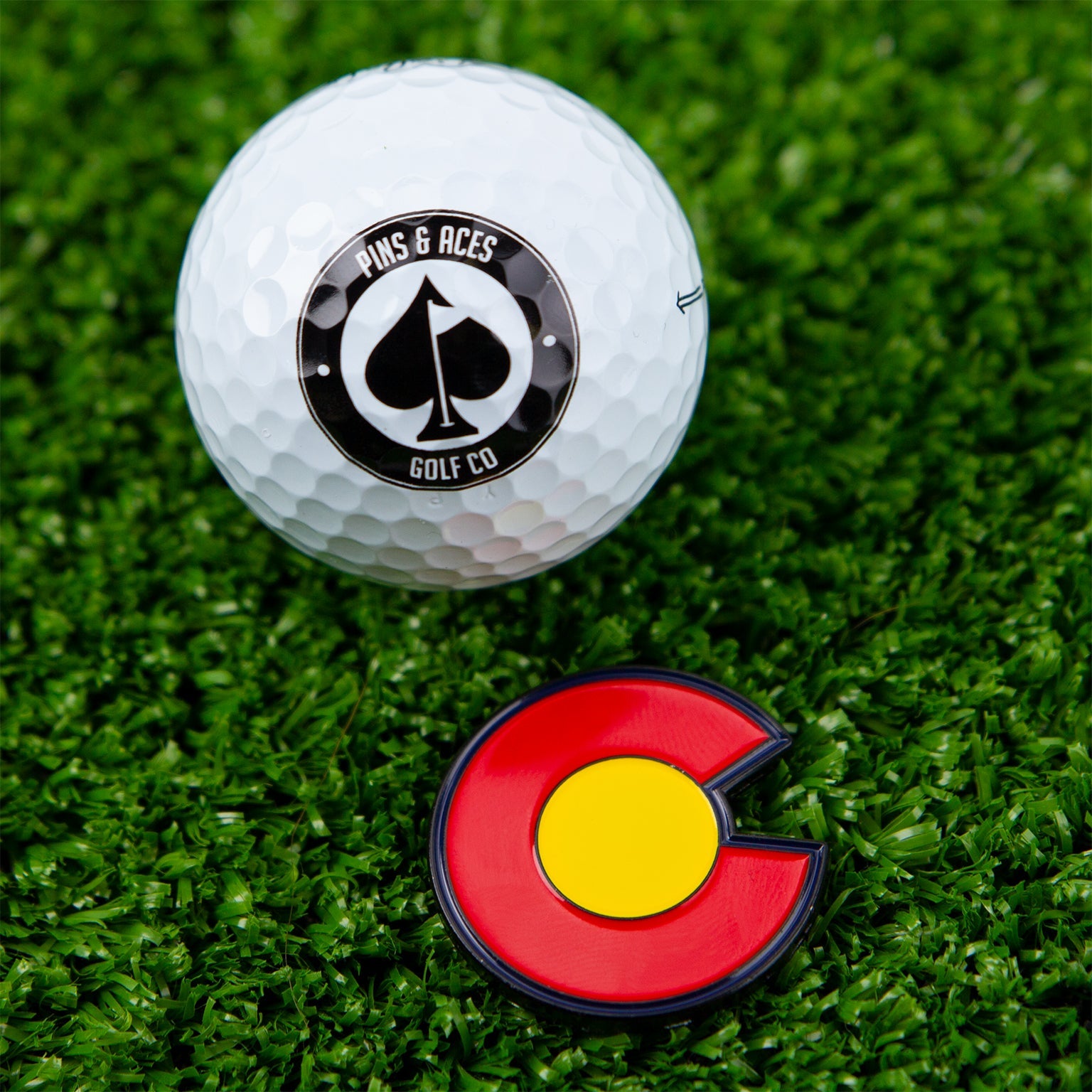 A Pins and Aces golf ball rests on green artificial grass beside the Colorado Strong Ball Marker, featuring a vibrant state flag design, securely attached to its magnetic base.
