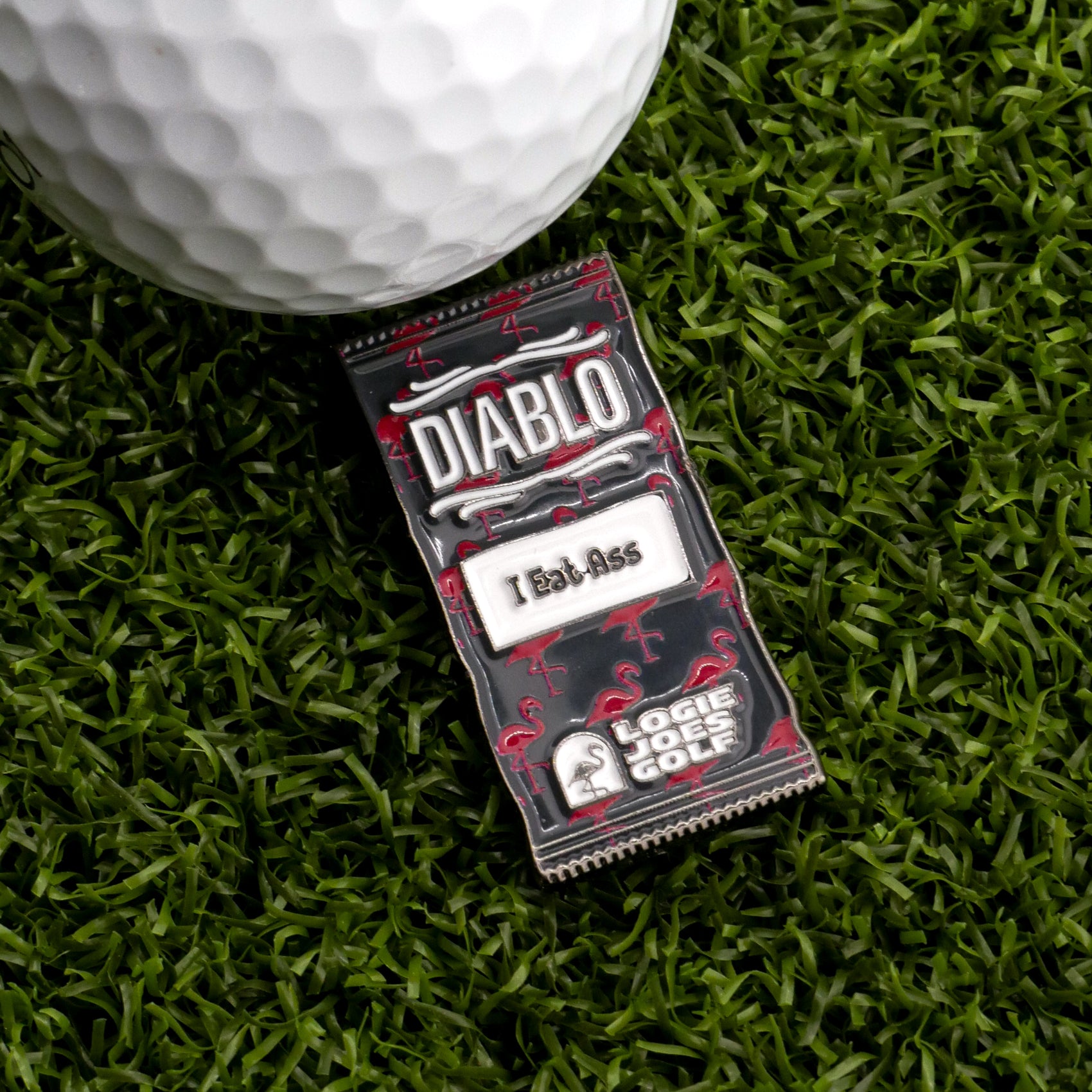 A close-up of a golf ball on artificial grass beside the Diablo Taco Sauce Packet Ball Marker, which humorously reads "DIABLO" and "I Eat Ass," featuring the Logie Joe's Golf logo at the bottom.