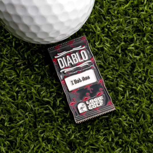A close-up of a golf ball on artificial grass beside the Diablo Taco Sauce Packet Ball Marker, which humorously reads 