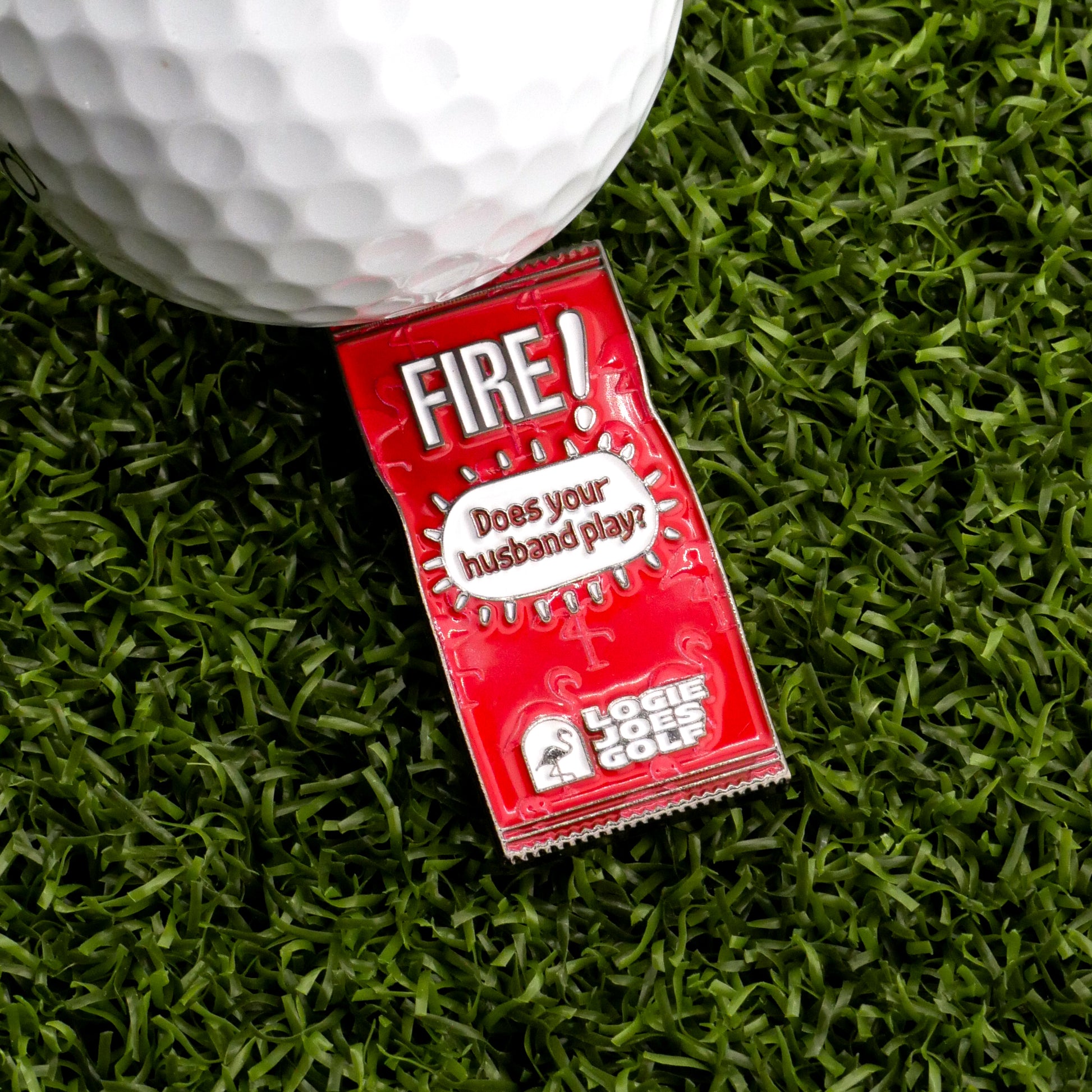 A white golf ball sits on artificial green grass beside a red Fire! Taco Sauce Packet Ball Marker from Logie Joe's Golf.