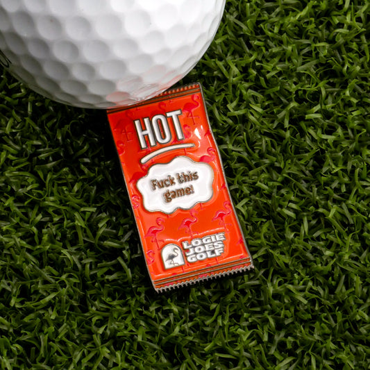 A close-up of a golf ball on artificial grass next to Logie Joe's Golf Hot Taco Sauce Packet Ball Marker, featuring 