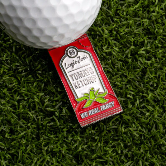 A golf ball rests on artificial grass next to the Logie Joe's Golf Tomato Ketchup Sauce Packet Ball Marker, featuring a pin with the phrase 