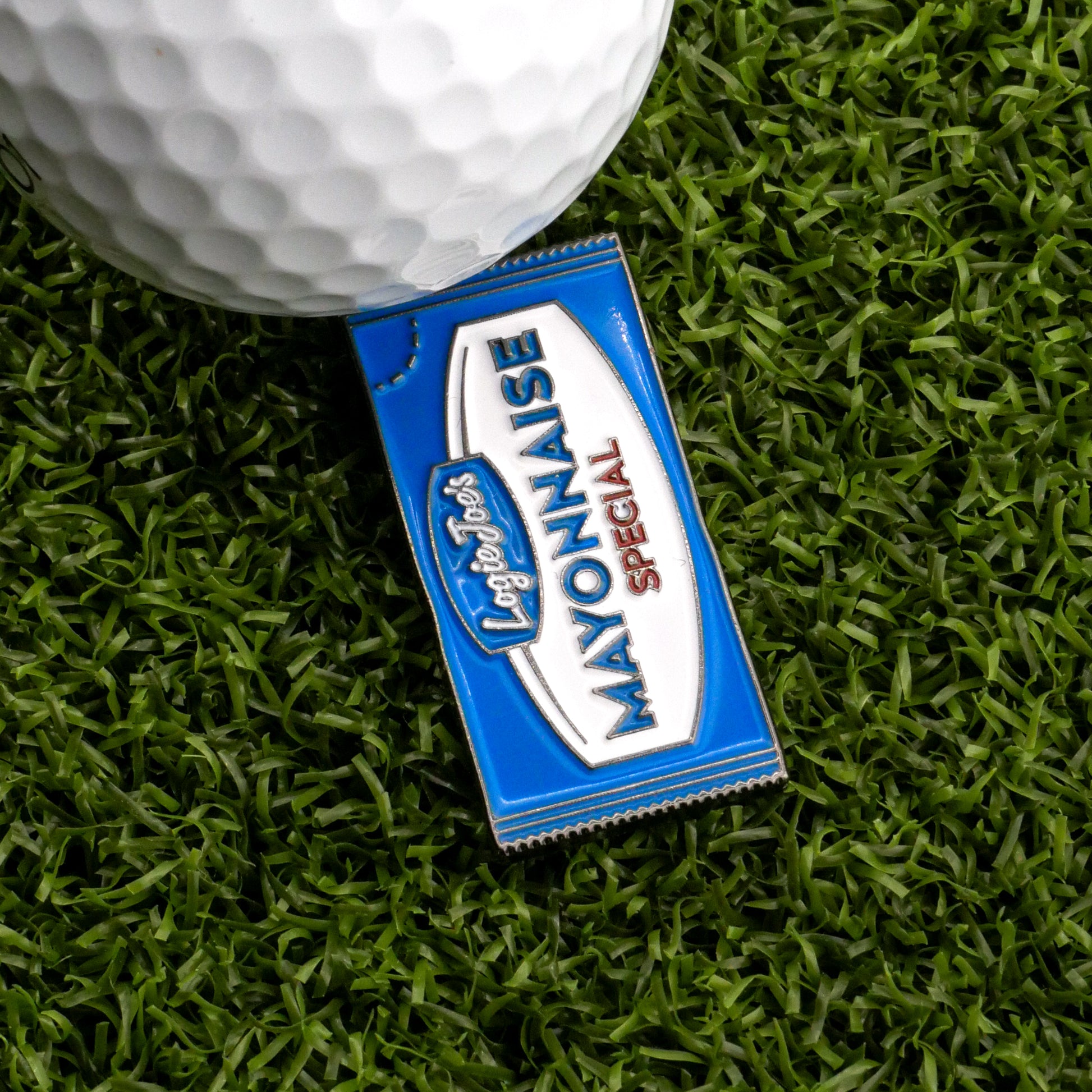 A blue and white Mayonnaise Sauce Packet Ball Marker from Logie Joe's Golf sits on green artificial grass next to a white golf ball.