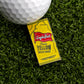 A golf ball sits on yellow artificial grass beside a Logie Joe’s Golf Yellow Mustard Sauce Packet Ball Marker, shaped like a mustard packet.