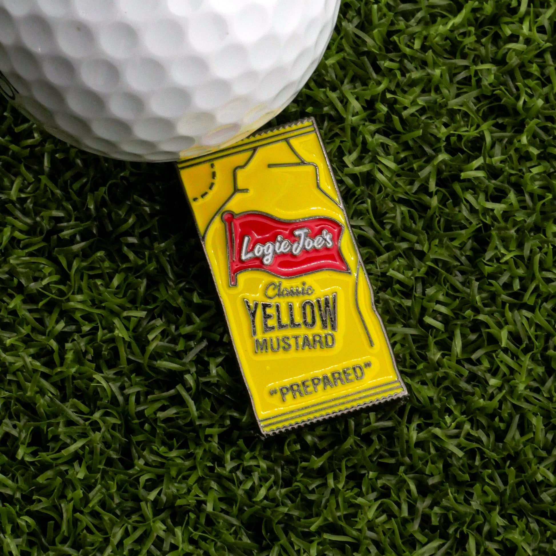 A golf ball sits on yellow artificial grass beside a Logie Joe’s Golf Yellow Mustard Sauce Packet Ball Marker, shaped like a mustard packet.
