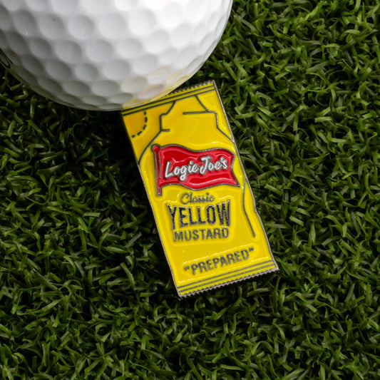 A golf ball sits on yellow artificial grass beside a Logie Joe’s Golf Yellow Mustard Sauce Packet Ball Marker, shaped like a mustard packet.