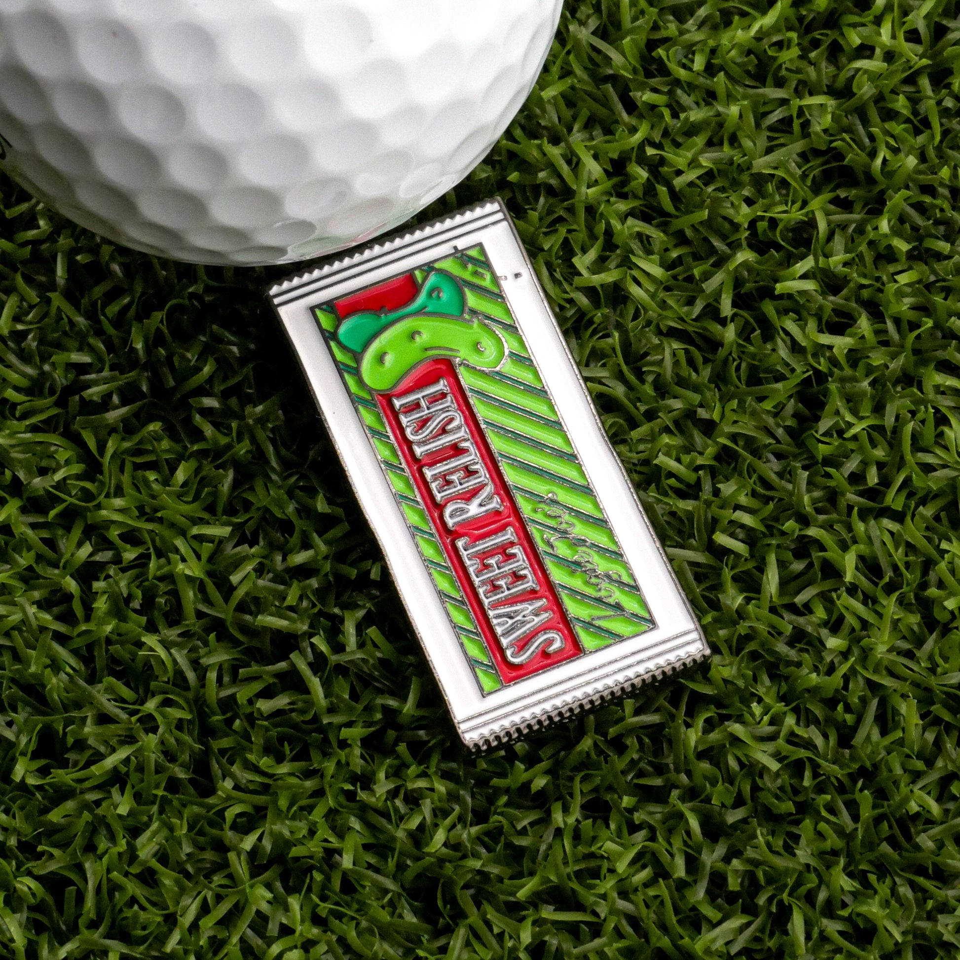The Logie Joe's Golf Sweet Relish Sauce Packet Ball Marker playfully mimics a relish packet with its colorful rectangular design and “Sweet Relief” print, shown beside a golf ball and green pin on artificial grass.