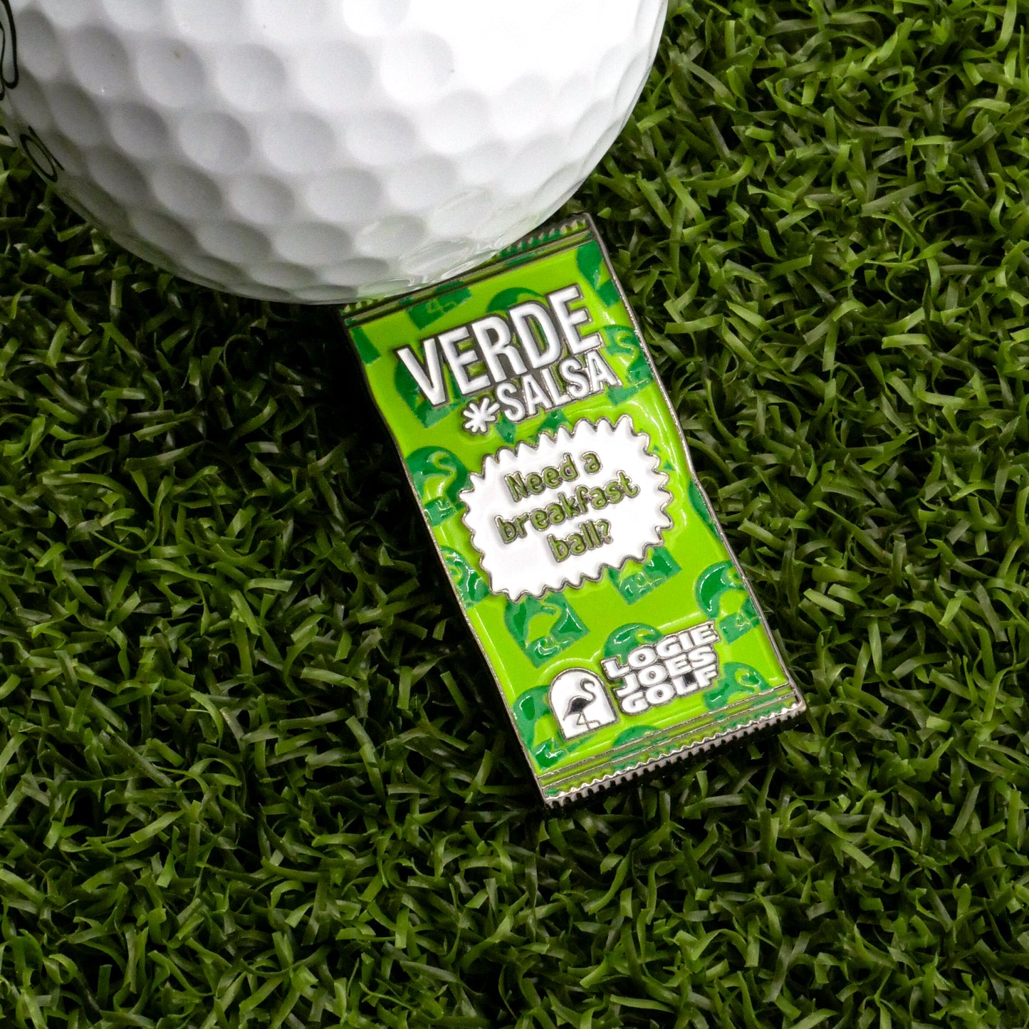 A Verde Salsa Taco Sauce Packet Ball Marker from Logie Joe's Golf, with golf-themed graphics, lies on artificial grass beside a white marker. The packet reads "Need a breakfast ball?" and displays the Logie Joe's Golf logo and a humorous message.