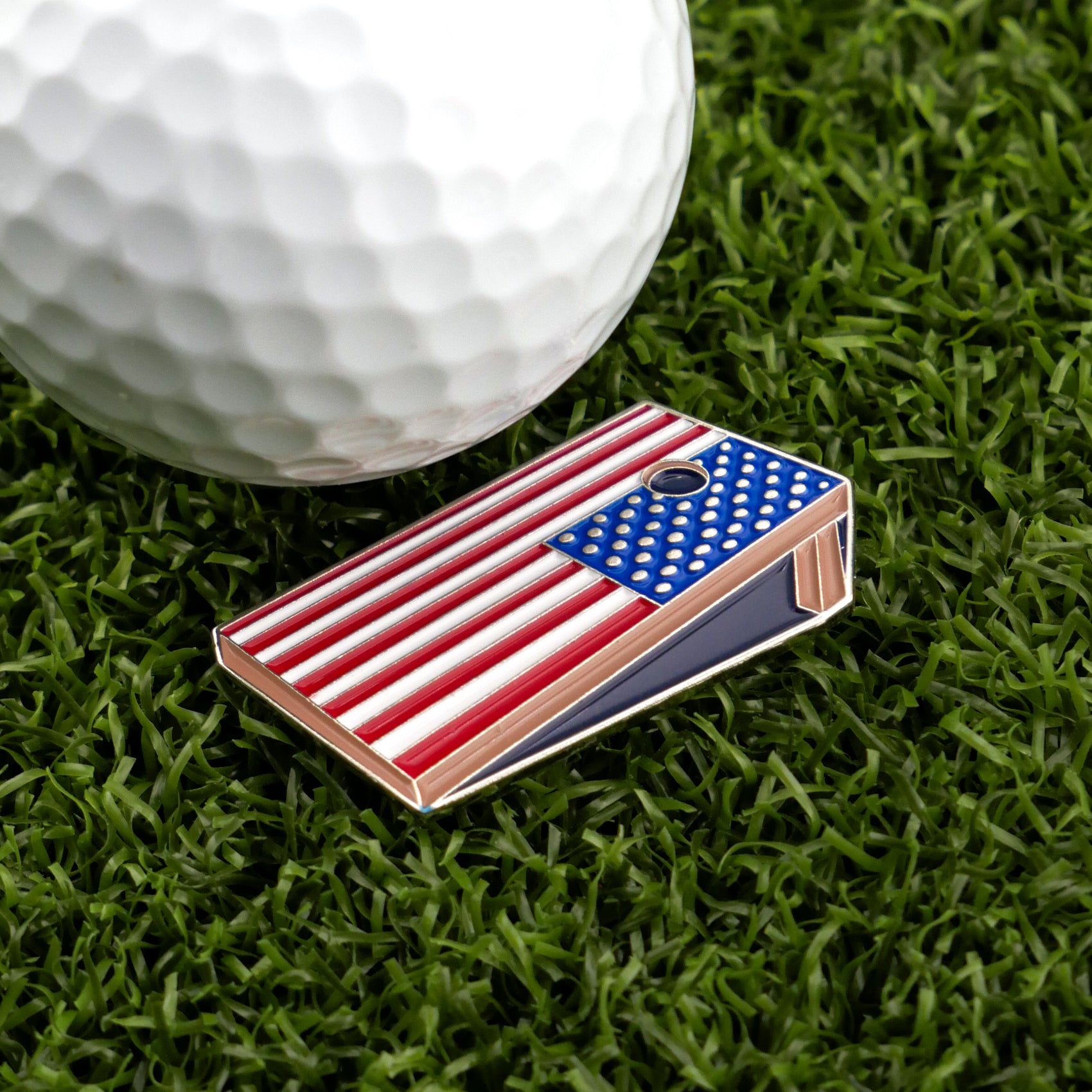 A golf ball sits on green grass beside Logie Joe's Golf Corn Hole Board, a mini cornhole board with an American flag design and circular hole—one of the funniest golf ball markers and great for funny golf gifts.