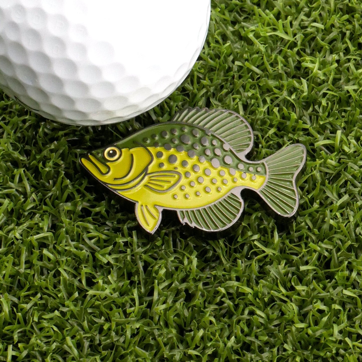 A golf ball on artificial grass sits next to Logie Joe's Golf Crappie Ball Marker, a green and yellow fish-shaped marker with detailed scales and fins—ideal for golfers seeking unique, premium accessories.