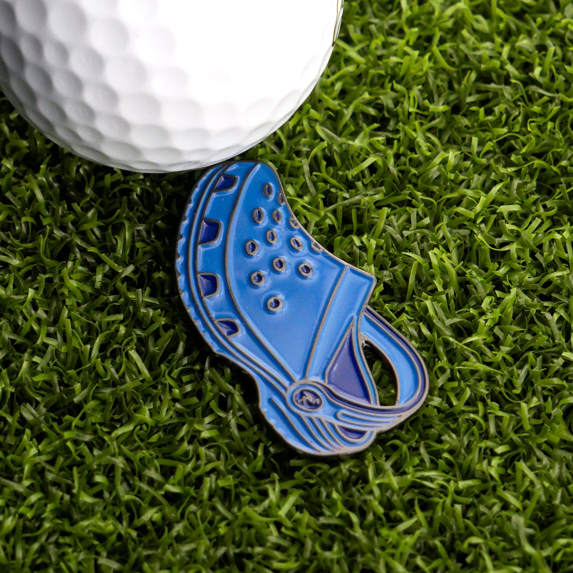 The Ballmarkers.com Rubber Clog Ball Marker, a fun blue golf accessory, sits on green artificial grass beside a white golf ball.