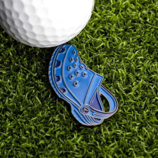 The Ballmarkers.com Rubber Clog Ball Marker, a fun blue golf accessory, sits on green artificial grass beside a white golf ball.