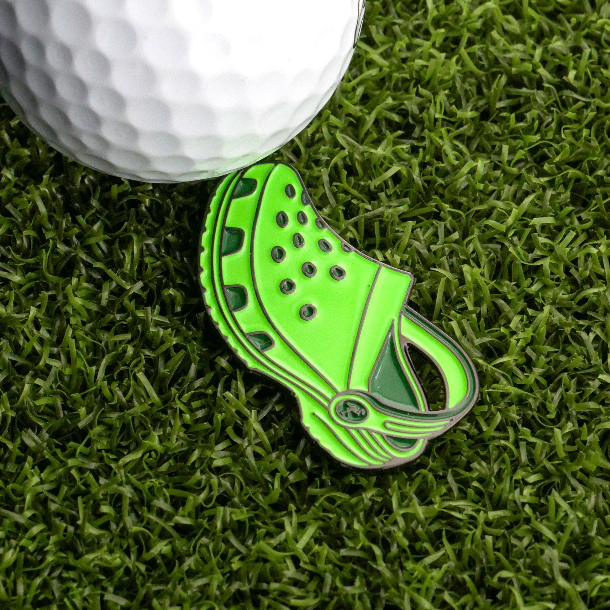 A white golf ball and the Rubber Clog Ball Marker from Ballmarkers.com, shaped like a green Crocs keychain, sit on artificial grass—a fun and unique golf accessory.