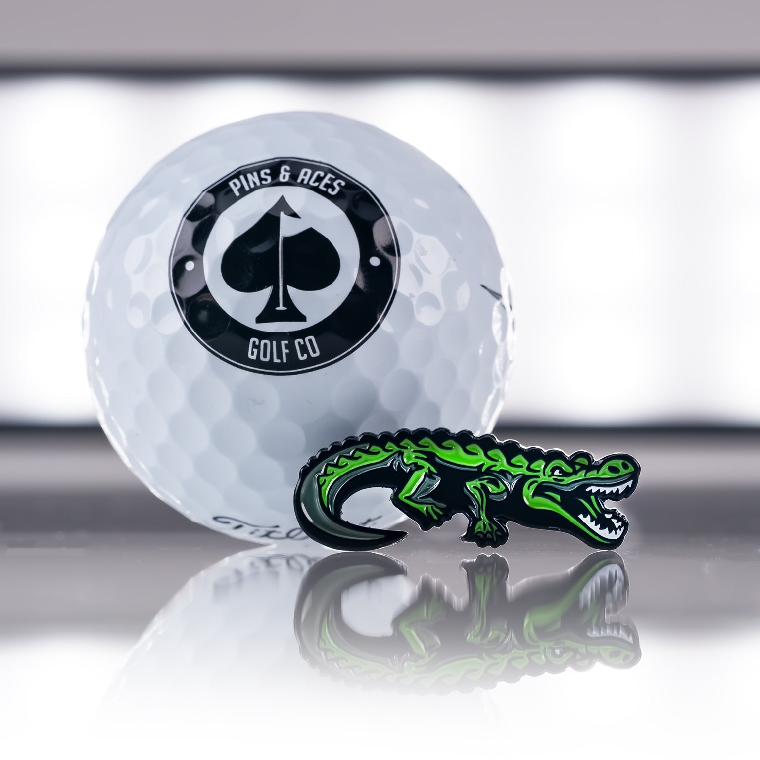 A white Pins and Aces golf ball stands beside the Crocodile Ball Marker— a small, green enamel marker shaped like a crocodile—both reflected on a shiny surface with a blurred background.