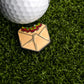 A golf ball sits on green artificial grass beside Logie Joe's Golf Crunchy Taco Wrap Ball Marker—an enamel pin shaped like a beige envelope with red chili peppers, making a quirky taco-themed golf accessory or unique gift.