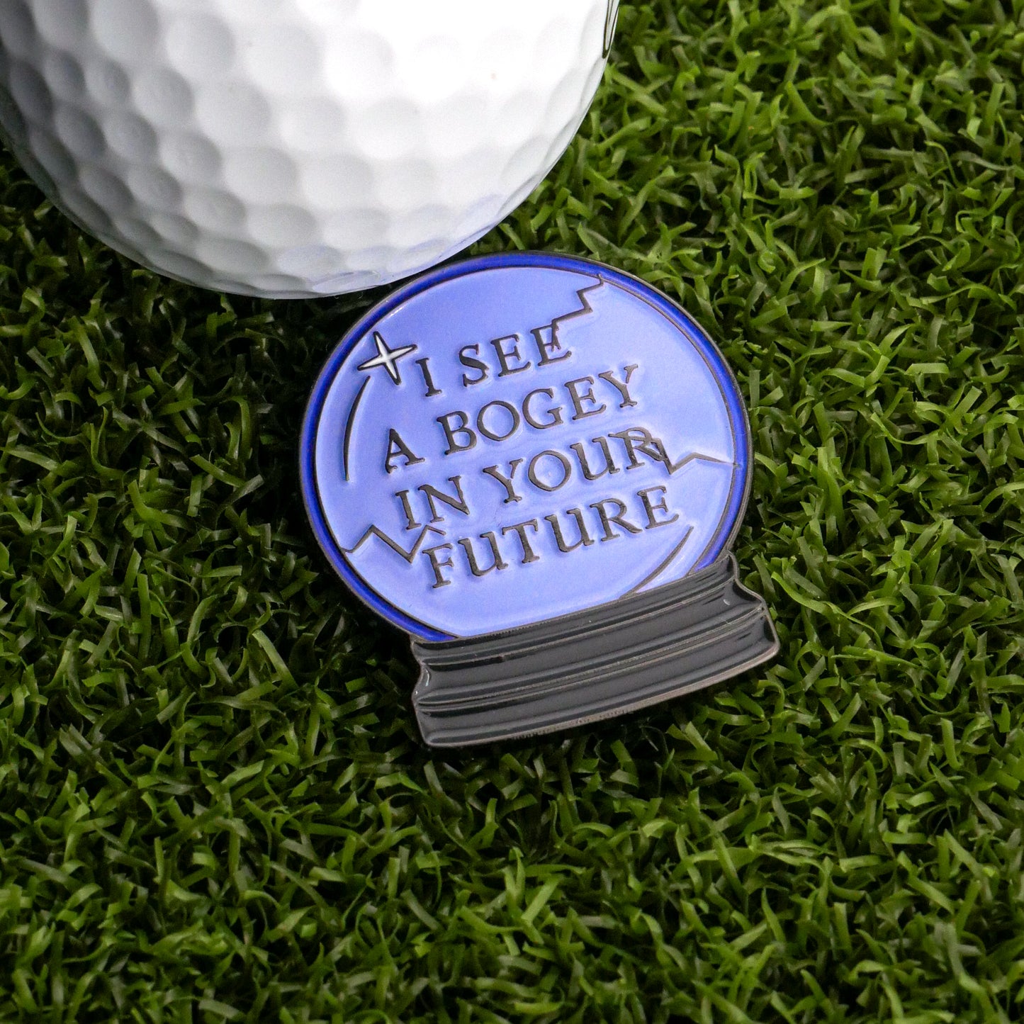 A golf ball sits on artificial grass next to Ballmarkers.com's I See A Bogey Crystal Ball Ball Marker, a blue orb warning, "I see a bogey in your future.