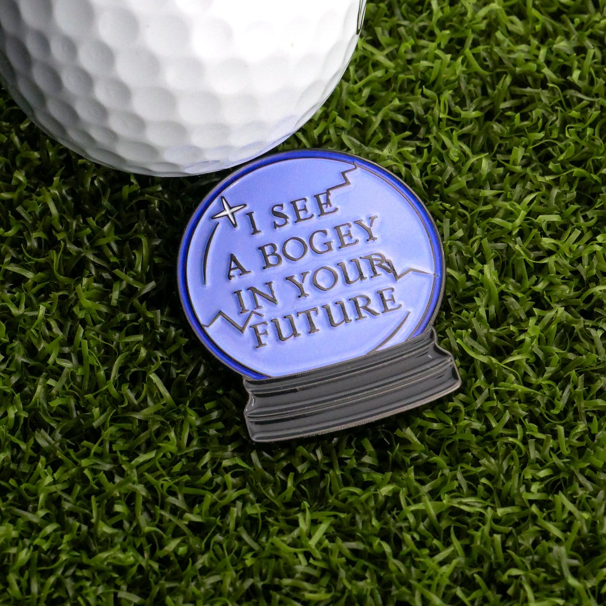 A golf ball sits on artificial grass next to Ballmarkers.com's I See A Bogey Crystal Ball Ball Marker, a blue orb warning, "I see a bogey in your future.