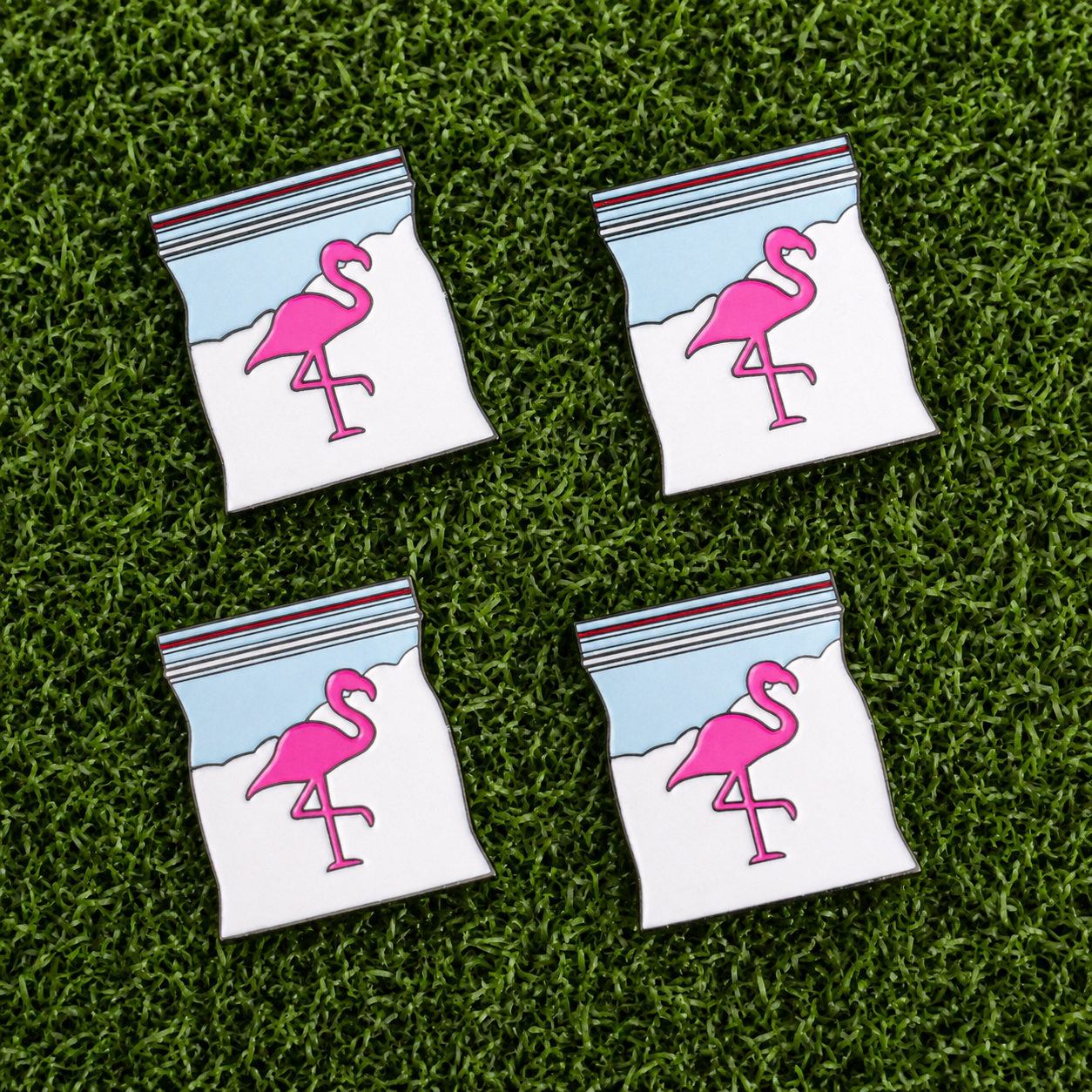 The Snowcane Baggie Ball Marker Bundle by Ballmarkers.com features four flamingo-themed pins, each shaped like a plastic bag with a blue, white, and red striped seal, arranged on green artificial grass.