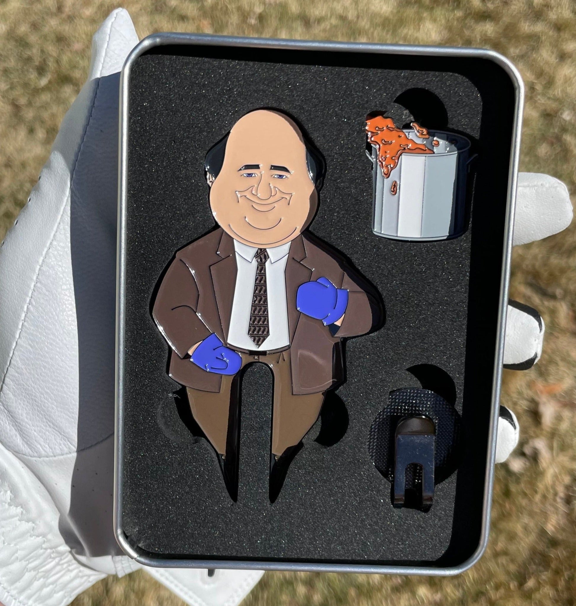 A gloved hand holds Pin Creatures’ Chili Cook Off Divot Tool set—a metal box with golf accessories in foam slots, featuring a cartoon bald man in a suit with blue gloves, a spilled chili pot, a black object, and a ball marker.