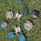 Shank it Golf's Blobett Ball Markers—colorful enamel pins with unique designs like skulls, creatures, and witty phrases—are displayed on green grass under bright sunlight.