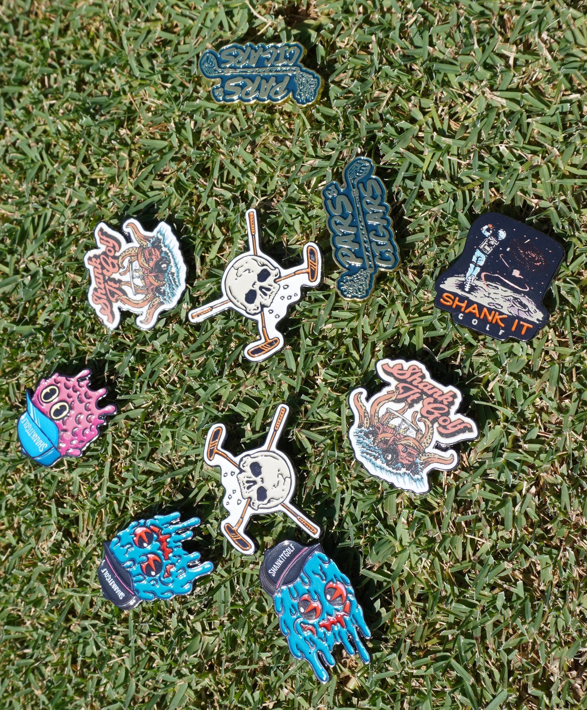 Shank it Golf's Blobett Ball Markers—colorful enamel pins with unique designs like skulls, creatures, and witty phrases—are displayed on green grass under bright sunlight.