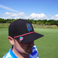 A man wearing a black hat with colorful embroidery stands on a golf course, sporting Shank it Golf's Blobett Ball Marker. The sky is blue with scattered clouds, and trees line the background as he looks down, partially facing away.