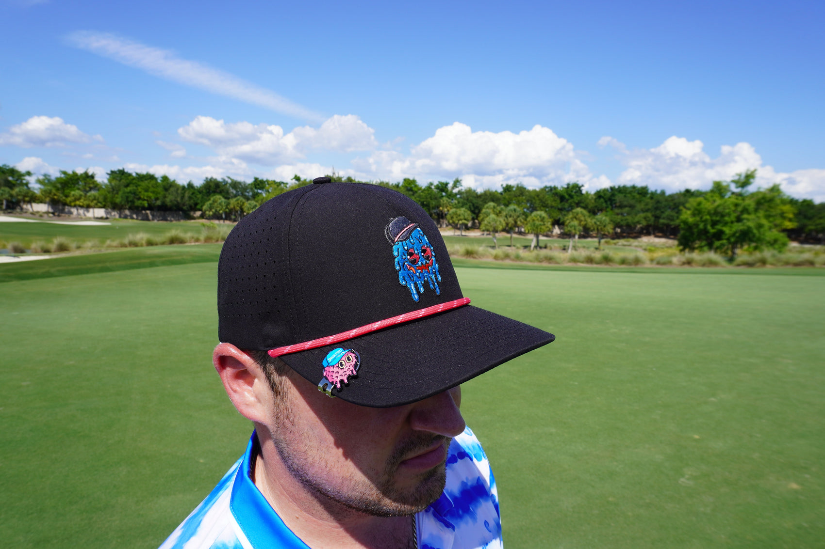 A man wearing a black hat with colorful embroidery stands on a golf course, sporting Shank it Golf's Blobett Ball Marker. The sky is blue with scattered clouds, and trees line the background as he looks down, partially facing away.