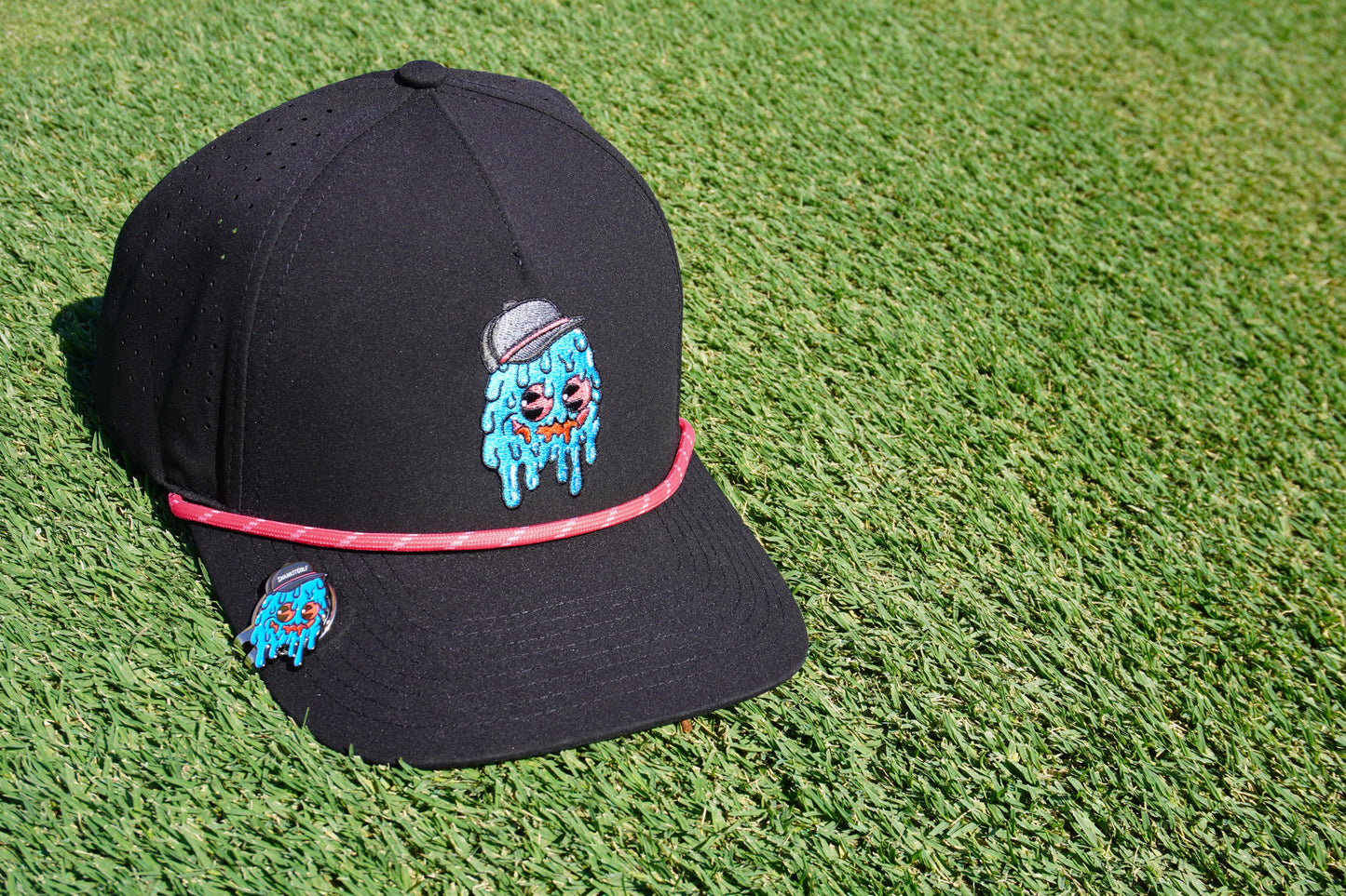 The Shank it Golf Blobert Ball Marker is a black cap featuring a colorful embroidered melting blue face in a gray cap, topped with a pink-brim band and premium metal magnetic ball markers, all displayed on green artificial grass.