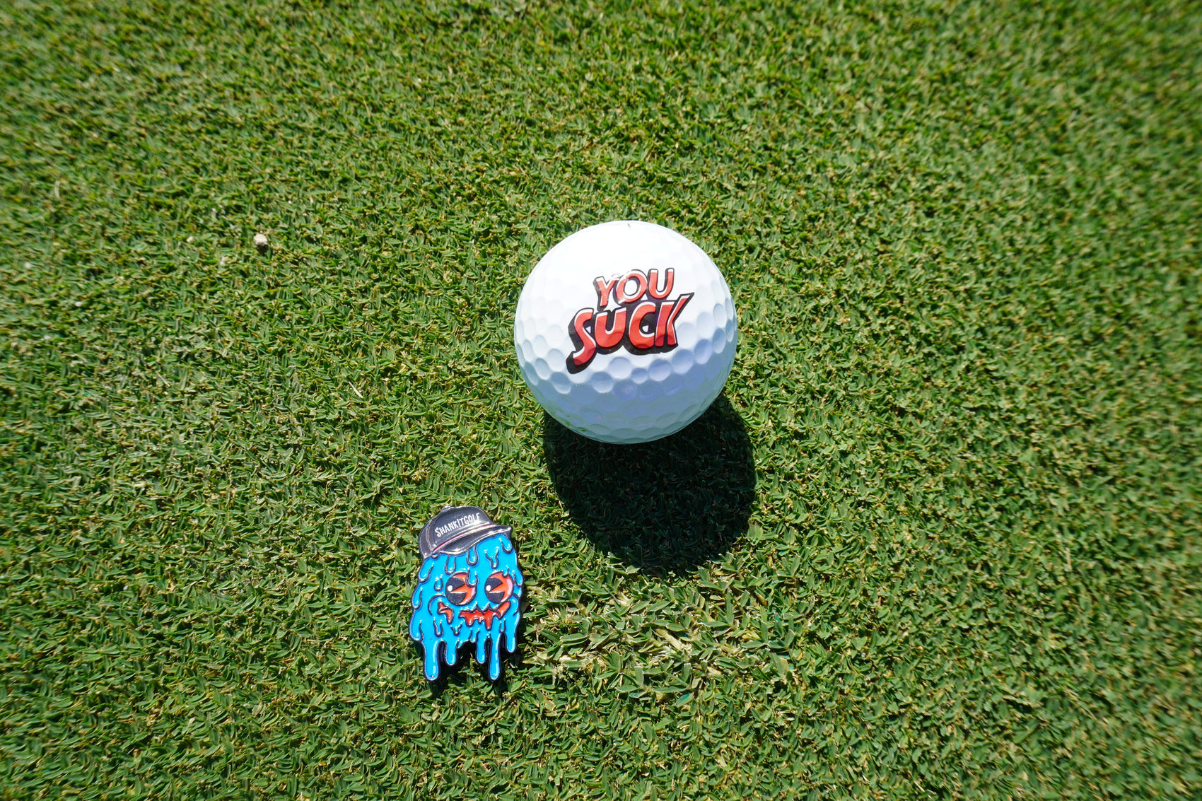A golf ball with "YOU SUCK" in red on green grass sits beside Shank it Golf's Blobert Ball Marker—a colorful, premium metal magnetic marker featuring a blue melting cartoon face in a gray cap.