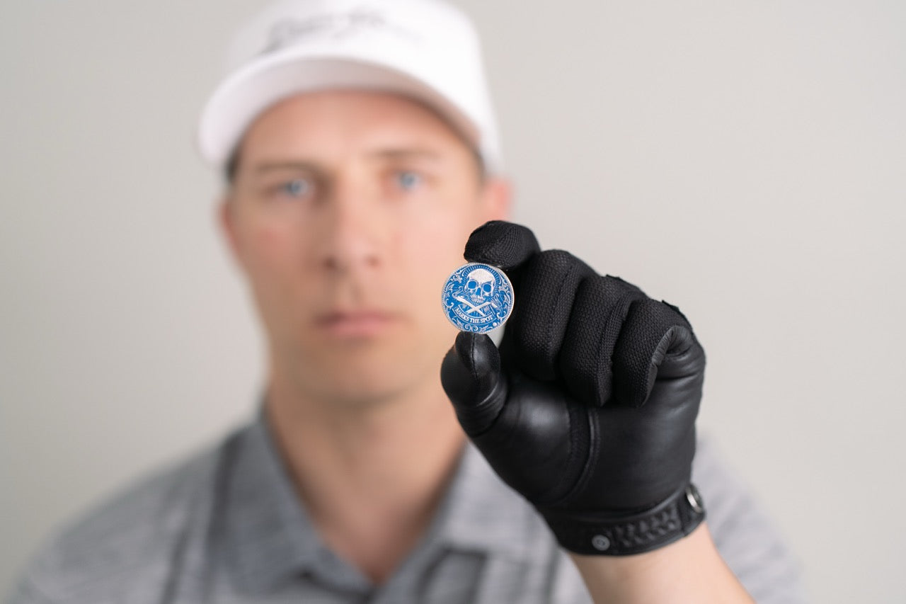 A person in a white cap and black glove holds Doubloon’s The Silver Omen Antique, Color Edition golf ball marker with a blue skull design, focusing on the marker while their face is blurred in the background.