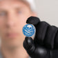 A person wearing a black glove holds Doubloon's The Silver Omen Antique, Color Edition—a blue and silver personalized golf ball marker with a skull design—while their blurred face is seen in the background.