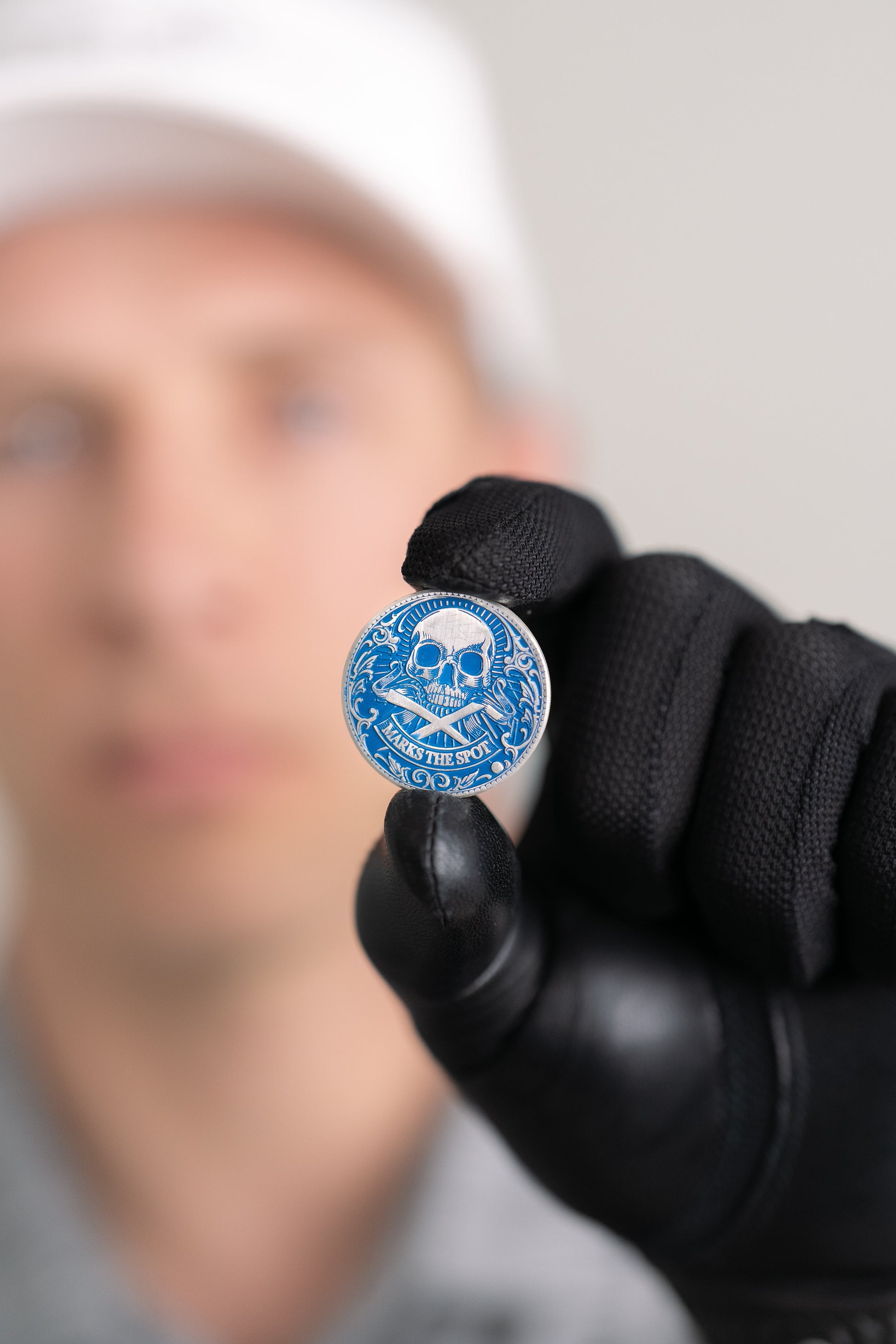 A person wearing a black glove holds Doubloon's The Silver Omen Antique, Color Edition—a blue and silver personalized golf ball marker with a skull design—while their blurred face is seen in the background.