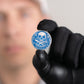 A person wearing black gloves holds Doubloon’s The Silver Omen Antique, Color Edition—a blue personalized golf ball marker featuring a skull and crossed swords with the words TAKE THE SOUL. The person's face is blurred in the background.