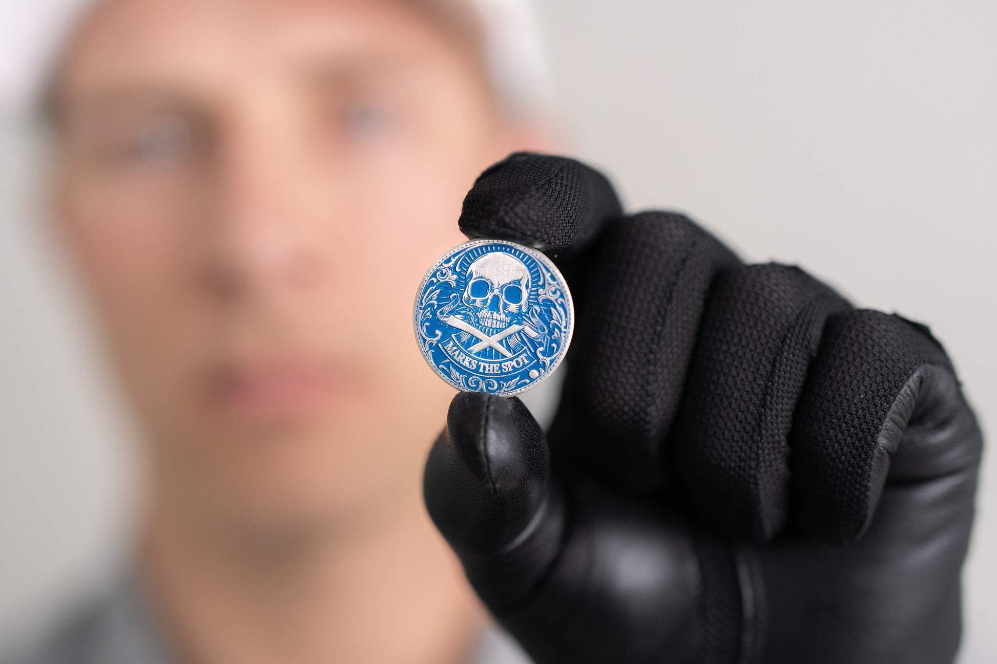 A person wearing black gloves holds Doubloon’s The Silver Omen Antique, Color Edition—a blue personalized golf ball marker featuring a skull and crossed swords with the words TAKE THE SOUL. The person's face is blurred in the background.