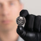 A black-gloved hand holds Doubloon’s The Omen Classic Antique, Diamond & Silver Edition golf ball marker, featuring a skull and crossbones with "X MARKS THE SPOT"; the person’s face appears blurred in the background.