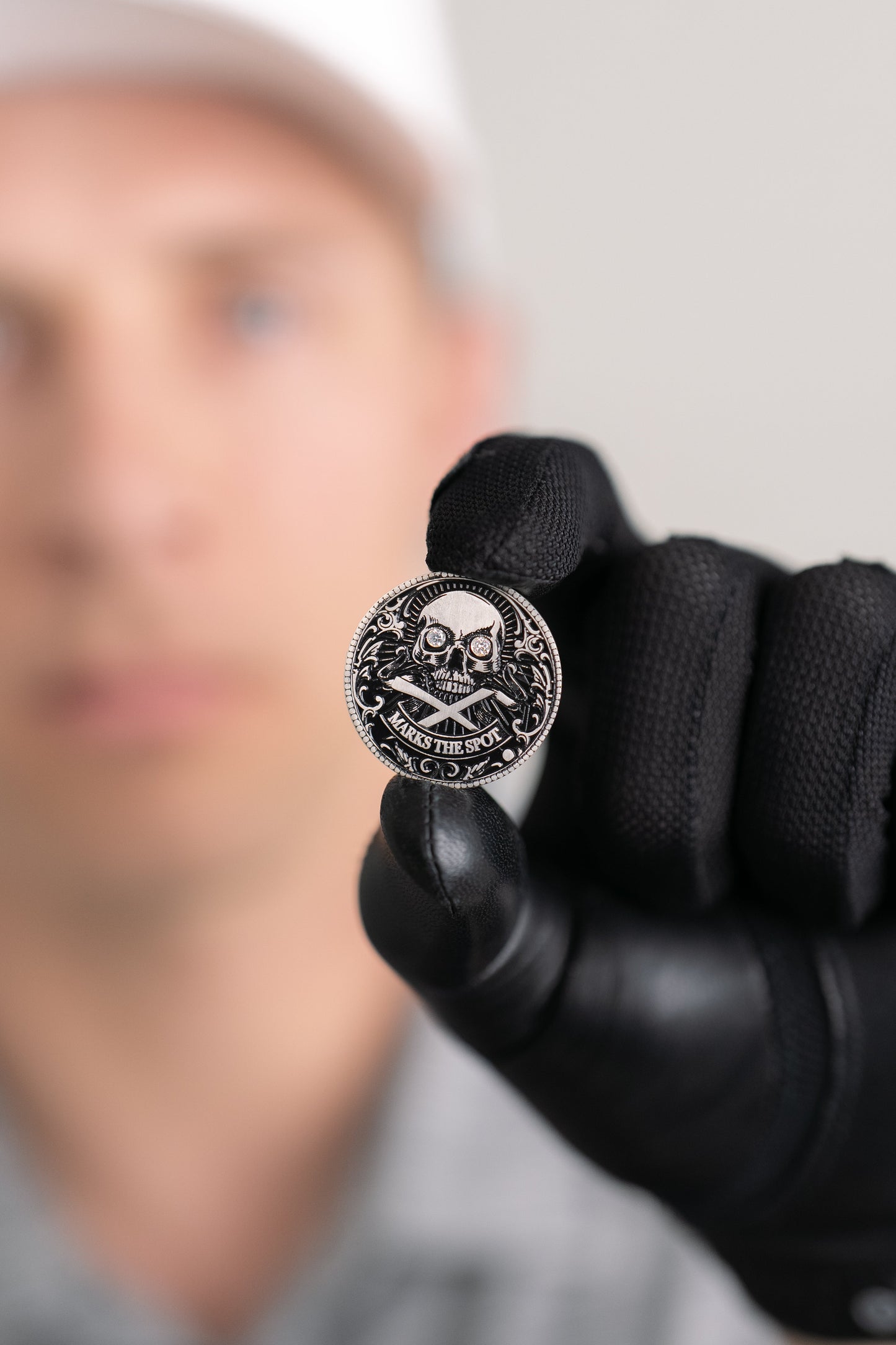 A black-gloved hand holds Doubloon’s The Omen Classic Antique, Diamond & Silver Edition golf ball marker, featuring a skull and crossbones with "X MARKS THE SPOT"; the person’s face appears blurred in the background.