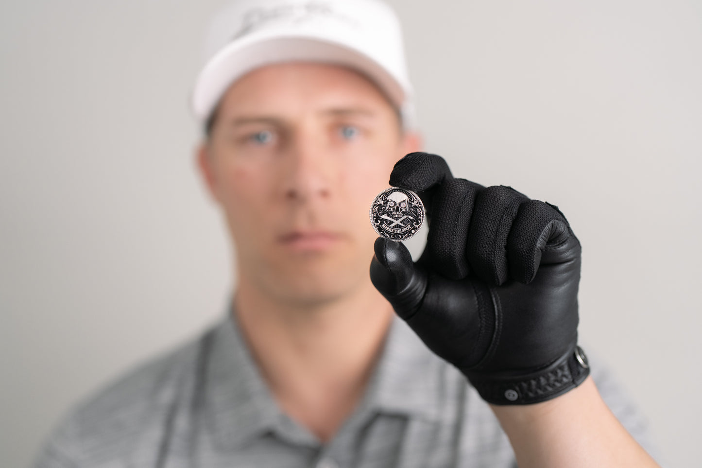 A person in a white cap and gray shirt, wearing a black glove, holds Doubloon’s The Omen Classic Antique, Diamond & Silver Edition golf ball marker toward the camera; their face is out of focus in the background.
