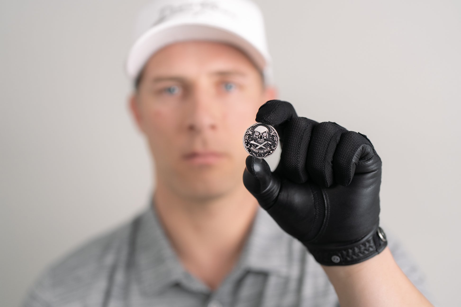 A person in a white cap and gray shirt, wearing a black glove, holds Doubloon’s The Omen Classic Antique, Diamond & Silver Edition golf ball marker toward the camera; their face is out of focus in the background.